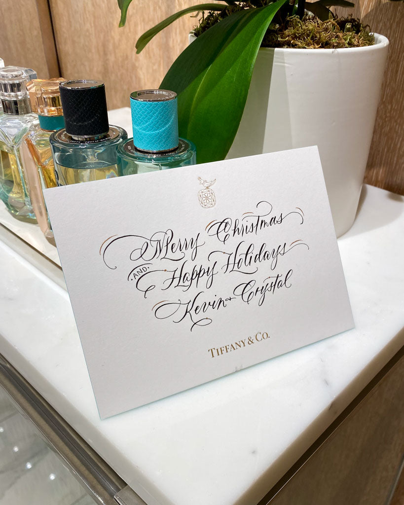 Elegant calligraphy holiday card reading “Merry Christmas and Happy Holidays” written for Tiffany & Co. Toronto, featuring fine script lettering beside fragrance bottles.