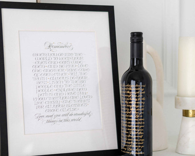 Calligraphy art in black frame and engraved wine bottle on white mantel display.