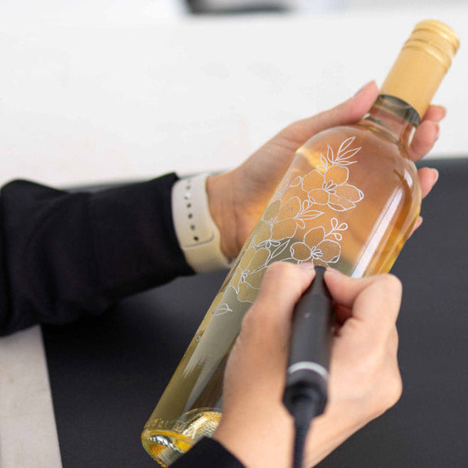 Toronto engraving artist creating intricate white floral design on wine bottle during live personalization event.